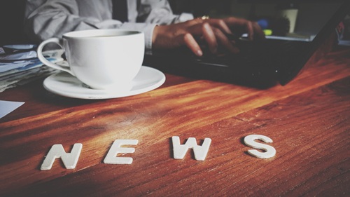 A person types on a computer with a cup of coffee and cut off letters that spell, "news" sit on the desk.typing on their computer at